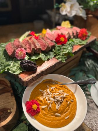 Farm-to-table presentation: sliced medium-rare steak on leafy greens and a rustic wooden board with edible flowers, paired with a bowl of creamy orange dip topped with slivered almonds.