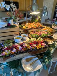 Vibrant catered buffet on a kitchen island with wooden boards of roasted chicken and sliced steak, a colorful heirloom tomato and watermelon-radish salad, bowls of dressings, gold serving utensils, decorative flowers and heat lamps.