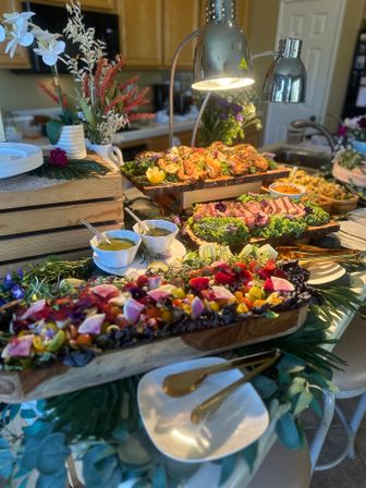 Vibrant catered buffet on a kitchen island with wooden boards of roasted chicken and sliced steak, a colorful heirloom tomato and watermelon-radish salad, bowls of dressings, gold serving utensils, decorative flowers and heat lamps.