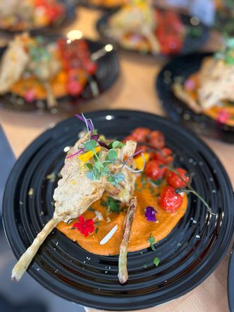 Tempura-style flower fritter skewers on smoky red pepper purée with blistered cherry tomatoes, edible flowers and microgreens served on a black plate
