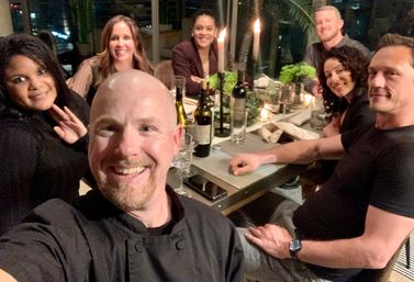 Smiling group of friends taking a selfie at a candlelit rooftop dinner table with wine bottles, glasses, greenery accents and a city skyline visible through windows at night