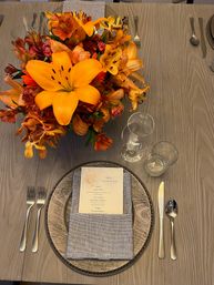 Autumn tablescape on gray wood dining table with vibrant orange lily centerpiece, round wooden charger, folded linen napkin topped by a printed menu card, wine and water glasses, and polished silver flatware.