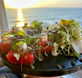 Bright watermelon-and-feta salad with microgreens, mint and frisée on a black plate, set against a golden ocean sunset view