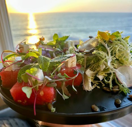 Bright watermelon-and-feta salad with microgreens, mint and frisée on a black plate, set against a golden ocean sunset view