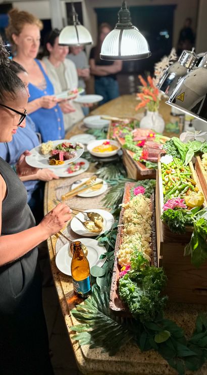 Guests at a lively indoor dinner party serving themselves from a well-lit buffet on a kitchen island with wooden trays of salads, roasted vegetables, rice, garnishes and heat lamps overhead.