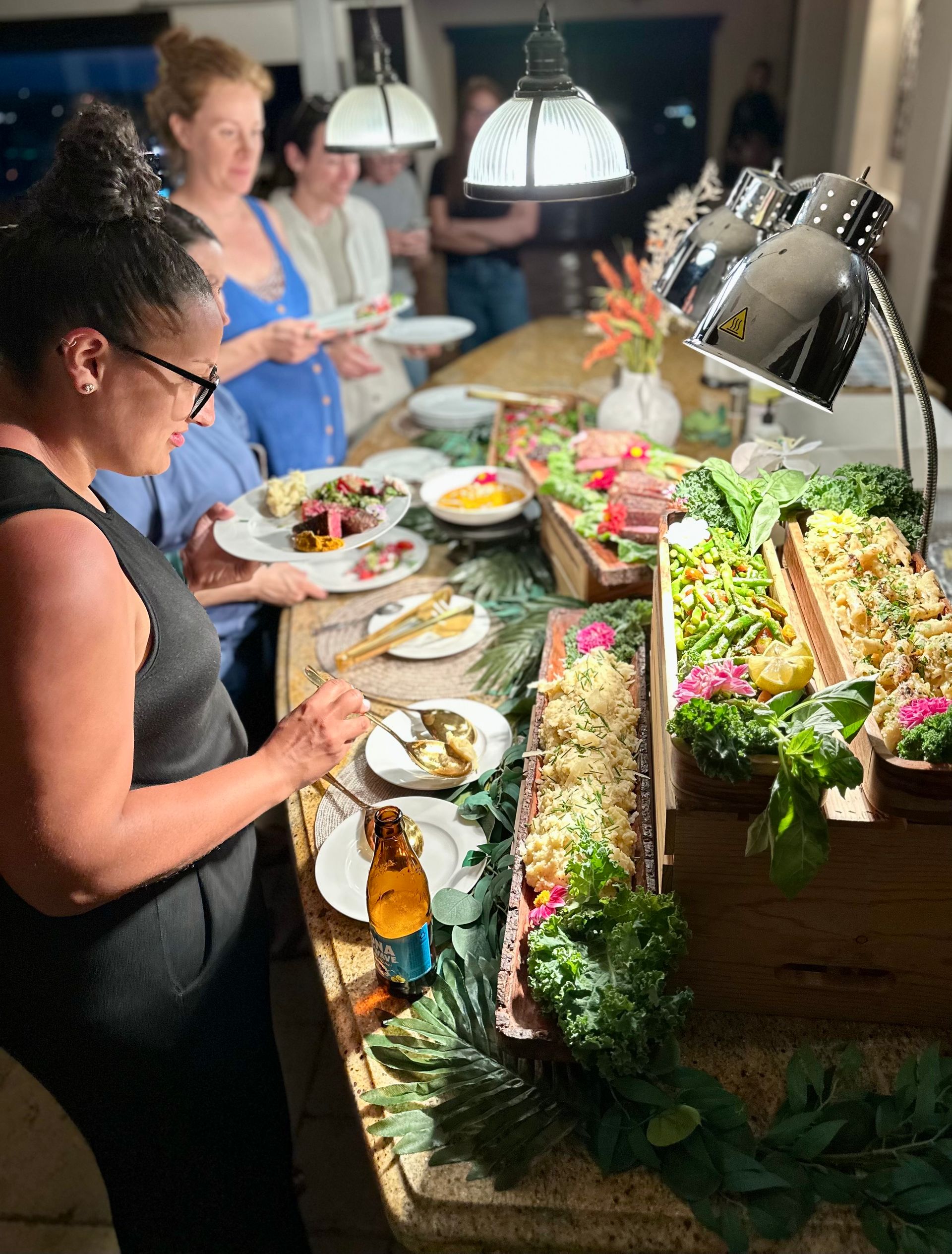 Guests at a lively indoor dinner party serving themselves from a well-lit buffet on a kitchen island with wooden trays of salads, roasted vegetables, rice, garnishes and heat lamps overhead.
