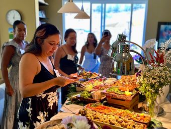Guest serving herself at an indoor catered buffet—heat lamps over platters of roasted vegetables, salads and seafood, bright floral arrangement and other guests waiting in line.