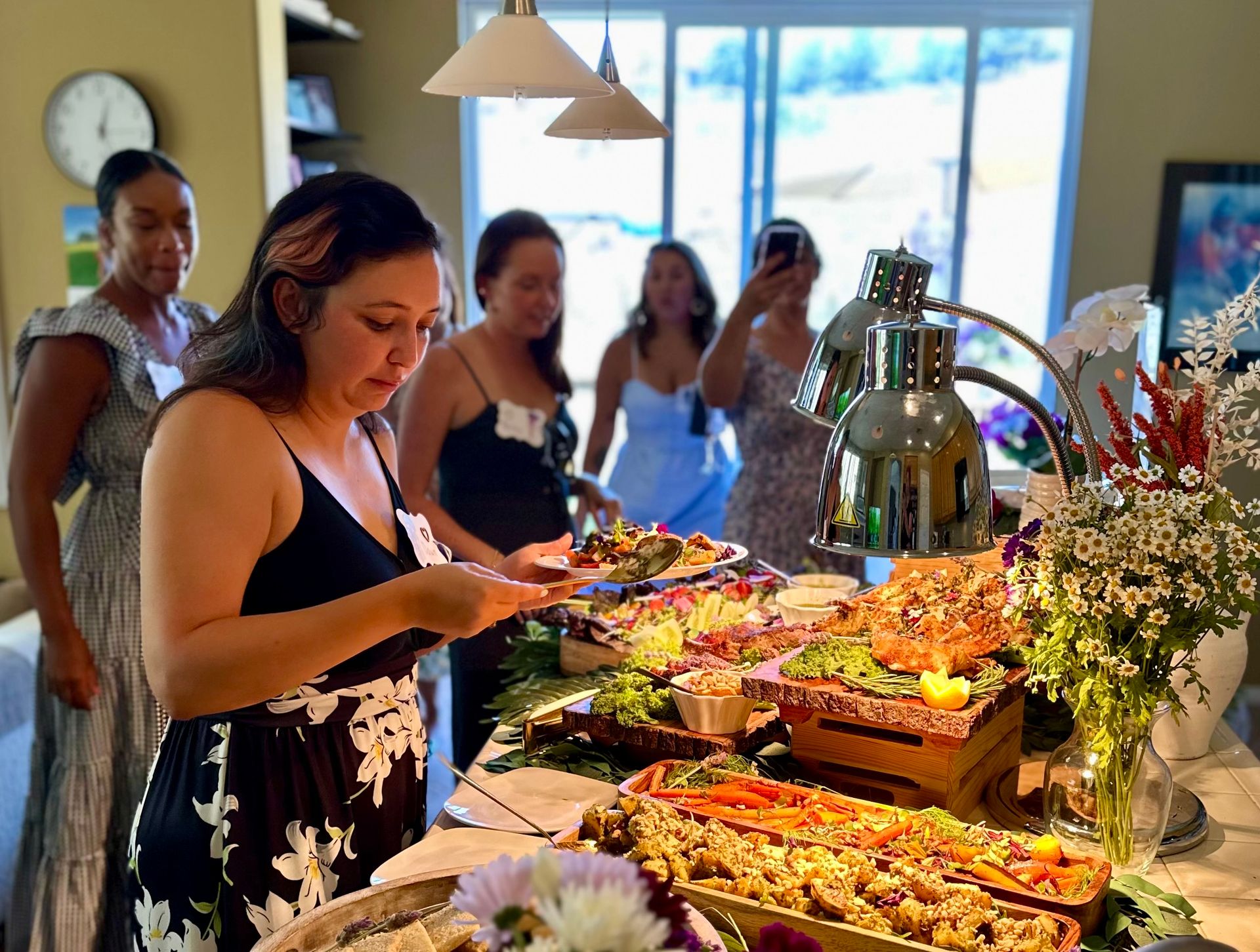 Guest serving herself at an indoor catered buffet—heat lamps over platters of roasted vegetables, salads and seafood, bright floral arrangement and other guests waiting in line.