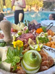 Close-up of a colorful charcuterie board at a poolside summer gathering: sliced pear, fresh figs, grapes, cured meats, cheeses, dried apricots, herbs and edible flowers with a wooden-handled cheese knife and blurred people and pool in the background.