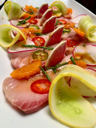 Bright sashimi-style citrus crudo appetizer on a white plate: thin raw fish slices topped with mandarin segments, red chili and jalapeño rounds, watermelon radish wedges, curled cucumber ribbons and green sea beans, drizzled with citrus dressing.