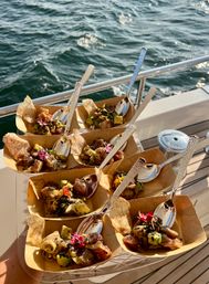 Sunlit mini poke seafood appetizers with avocado, diced tuna, sesame seeds and edible flowers in paper trays with spoons, served on a boat deck with ocean waves and yacht railing in the background.