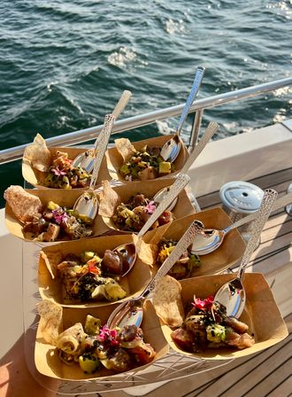 Sunlit boat-deck serving of tuna poke appetizers with avocado, sesame, edible flowers and taro chips in small paper trays with spoons, overlooking sparkling ocean waves.