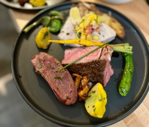 Gourmet restaurant plate with pink medium-rare roast, sliced cured sausage, chanterelle mushrooms, grilled zucchini and yellow squash, baby carrot and herb garnish on a black plate.