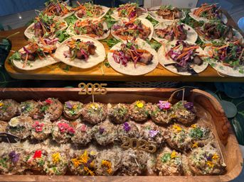 Colorful catering spread on wooden trays: mini tacos with grilled protein, shredded carrots, purple cabbage and microgreens on small tortillas, and stuffed mushrooms topped with grated cheese, herbs and edible flowers, accented by gold "2025" picks.