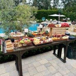 Backyard poolside grazing table on a stone patio with wooden platters of charcuterie, assorted cheeses, crackers, sliced fruit and grapes, prosciutto skewers and candles beside a blue swimming pool and lounge umbrellas.