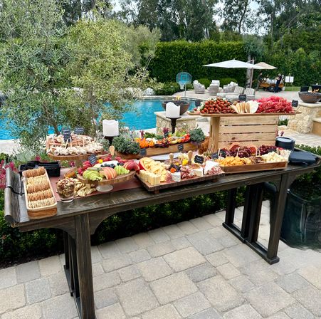 Poolside outdoor grazing table on a patio with wooden boards of charcuterie, assorted cheeses, crackers, fresh fruit, skewers and nuts, set against a backyard pool and lush greenery.