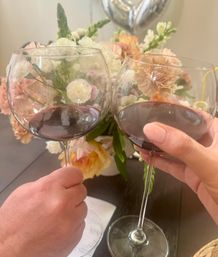 Two hands clinking red wine glasses over a pastel floral centerpiece on a home dining table — cozy indoor toast