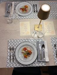 Overhead view of a modern two-person dining table set with plated citrus and radish salad, wine and water glasses, printed menus, silver flatware on blue-and-white patterned placemats and a small gold table lamp — cozy restaurant-style setting.