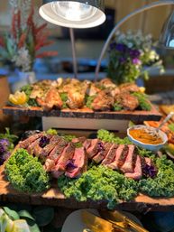 Carving station with sliced medium-rare steak on a rustic wooden board, garnished with curly kale and purple edible flowers, roasted chicken on a raised board behind, warm heat lamp overhead and a bowl of savory sauce in a buffet-style display.