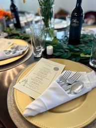 Elegant reception place setting: gold charger plate with folded white napkin holding forks and spoon, printed menu card, textured glassware, greenery table runner, votive candle and a wine bottle blurred in the background.