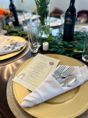 Elegant reception place setting: gold charger plate with folded white napkin holding forks and spoon, printed menu card, textured glassware, greenery table runner, votive candle and a wine bottle blurred in the background.