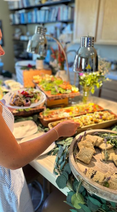 Guest serving from a garden-inspired appetizer buffet on a home kitchen island — trays of salads, roasted vegetables, and rustic crackers on a wooden platter under heat lamps.