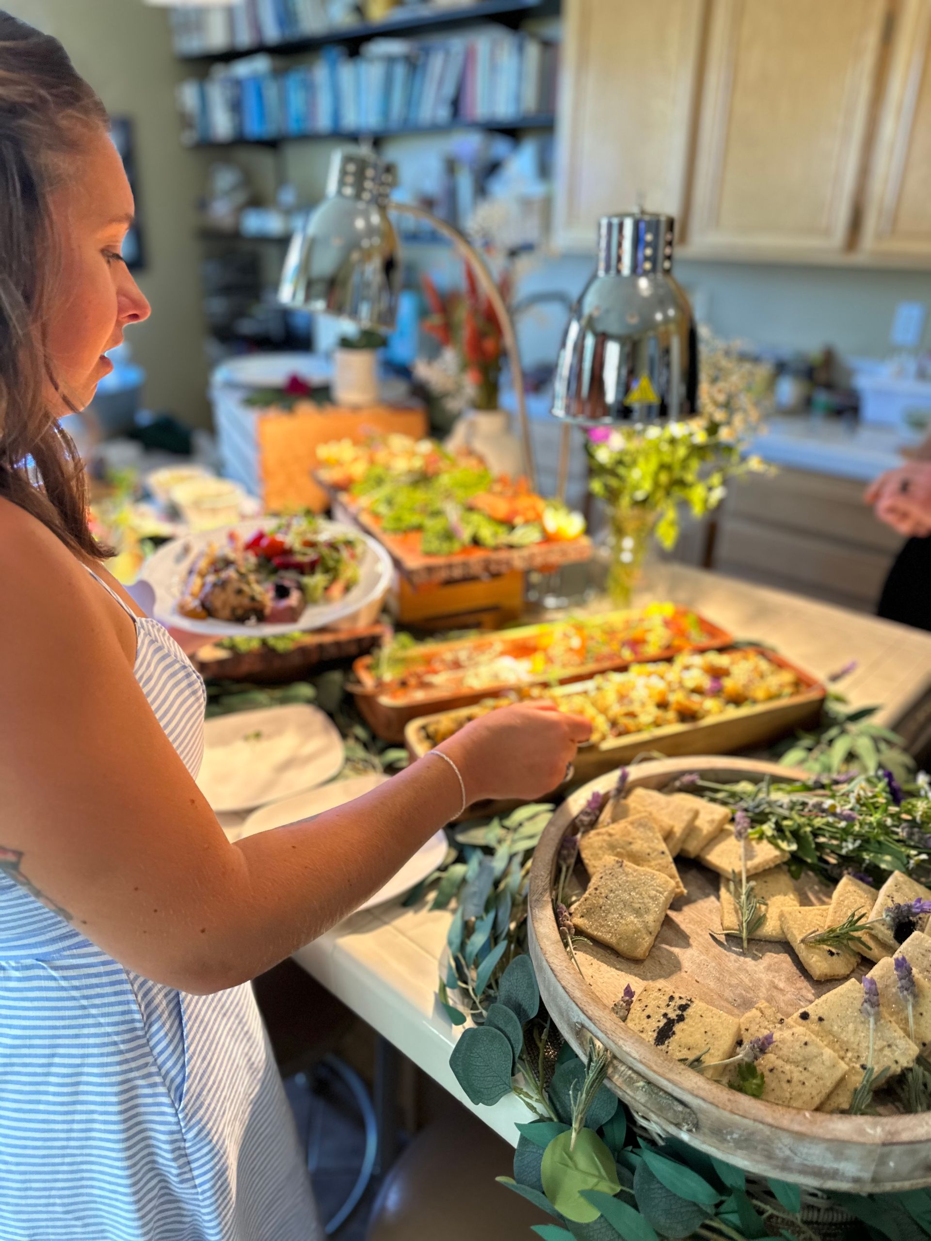 Guest serving from a garden-inspired appetizer buffet on a home kitchen island — trays of salads, roasted vegetables, and rustic crackers on a wooden platter under heat lamps.