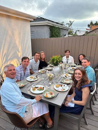 Eight people enjoying an al fresco dinner on a backyard patio at dusk — long table with plated summer dishes, wine glasses, a floral centerpiece, string lights and palm trees behind a wooden fence.