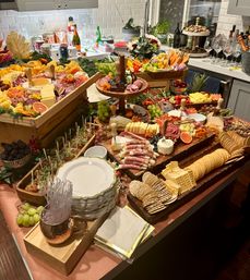 Festive grazing table on a kitchen island featuring layered charcuterie and cheese boards, assorted crackers, cured meats, grapes and berries, fresh fruit, veggies, nuts and stacked plates ready for a party