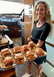 Smiling server on a luxury yacht holding a tray of mini pulled-pork sliders with pickled onions and slaw in a boat cabin — upscale yacht catering and party appetizers.