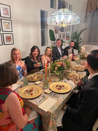 Candlelit dinner party in a cozy modern home dining room with guests seated around a table, gold charger plates holding plated appetizers, wine glasses, taper candles, floral centerpiece and glass-paned chandelier overhead.