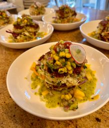 Crispy stacked crab cakes in a white bowl topped with corn-mango salsa, thin radish slice and green herb sauce — colorful seafood appetizer on a granite countertop.