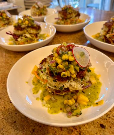 Crispy stacked crab cakes in a white bowl topped with corn-mango salsa, thin radish slice and green herb sauce — colorful seafood appetizer on a granite countertop.