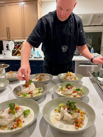 Chef in a black apron plating multiple bowls of white fish over rice with creamy sauce, romanesco florets, diced fruit salsa and fresh herbs on a home kitchen counter.