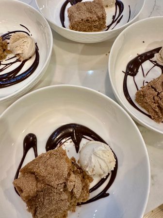 Overhead view of four white dessert bowls each with a scoop of vanilla ice cream and a piece of cinnamon crumb cake on a playful chocolate sauce swirl.