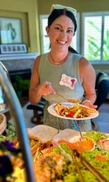 Smiling woman in a green tank top with sunglasses on her head ladling dressing onto a vibrant salad from a buffet spread in a sunlit living room, wearing a decorative name tag.
