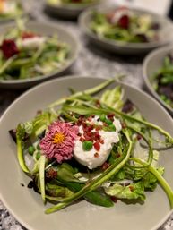 Close-up of a fresh spring salad with creamy burrata, sugar snap peas, shaved asparagus, mixed greens, crispy bacon bits and a pink edible flower on a gray plate.