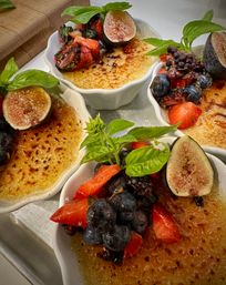 Crème brûlée in white ramekins with caramelized sugar tops, garnished with halved figs, blueberries, strawberries, blackberries and fresh basil on a tray — a vibrant plated dessert.
