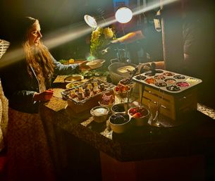 Evening dessert bar on a granite island at a party — guest reaching for a bowl under warm lamps beside trays of fruit, berries, marshmallows, chocolate toppings and crackers