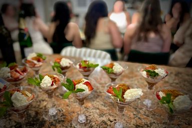 Row of strawberry parfaits with whipped cream, mint leaves and crumb topping served in martini glasses on a granite countertop, blurred guests gathered in the background at a party