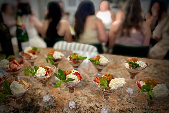 Row of strawberry parfaits with whipped cream, mint leaves and crumb topping served in martini glasses on a granite countertop, blurred guests gathered in the background at a party