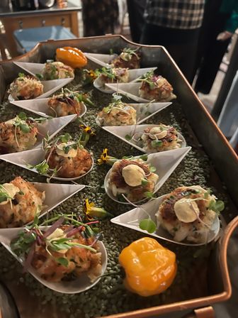 Catering tray of bite-sized seafood cakes on individual porcelain spoons, topped with aioli, microgreens and edible flowers, served as event appetizers with a bright yellow pepper garnish.