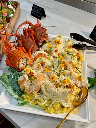Platter of cooked spiny lobsters and lobster meat on yellow seasoned rice, garnished with edible flowers and greens on a white serving tray at a buffet.