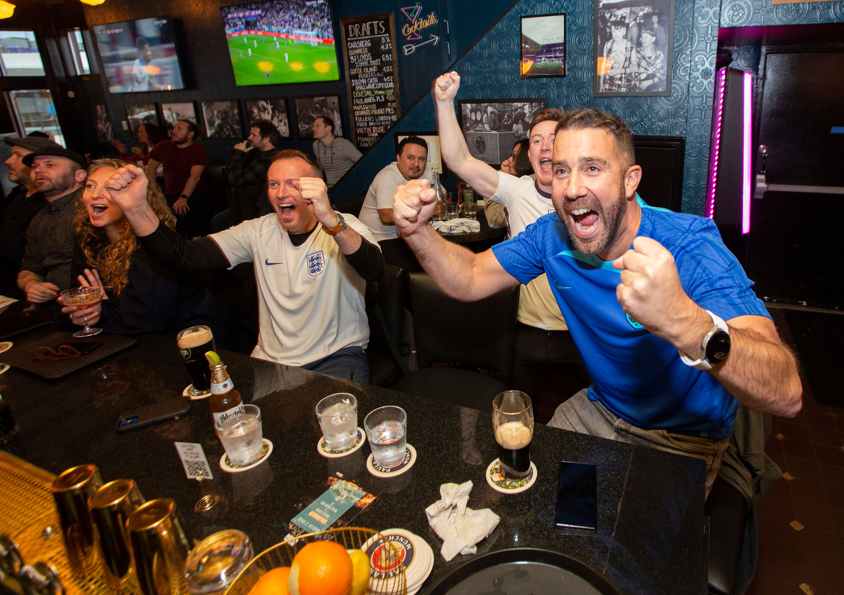 Lively sports bar crowd cheering with fists raised as fans in England shirts watch a soccer match on wall TVs, pints and glasses on the bar in a packed pub.