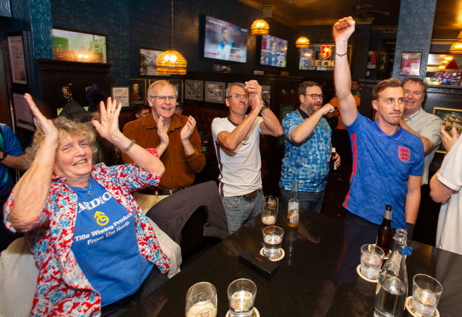 Pub crowd of football fans cheering and clapping with raised arms, wearing England shirts, bottles and glasses on the table, watching a match on TV screens in a cozy bar