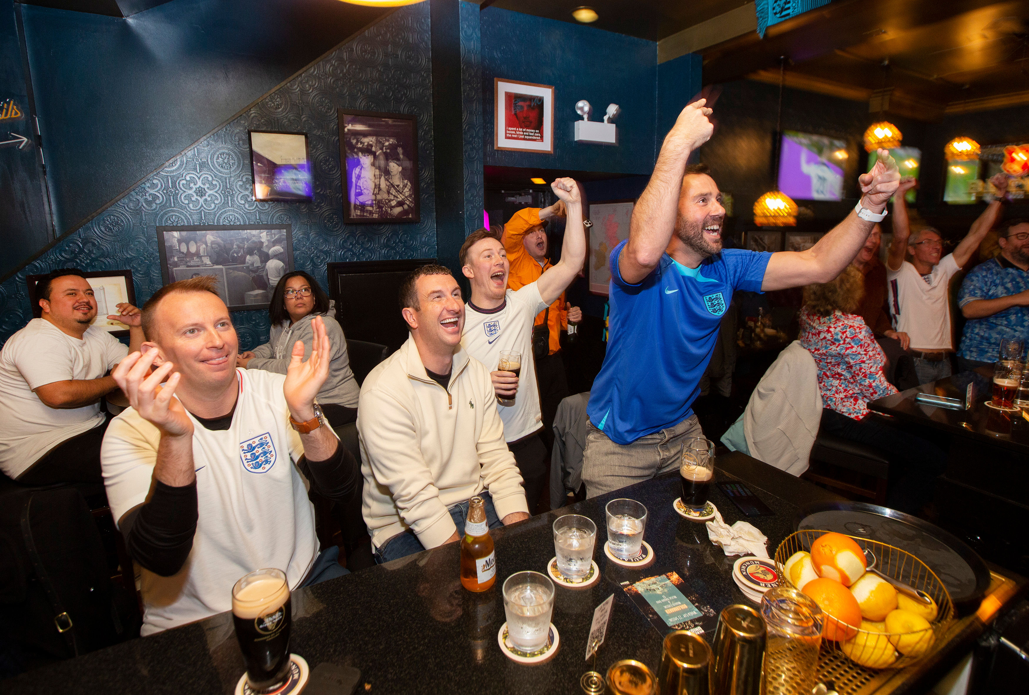 Cheering football fans at a lively pub watch party, many wearing England shirts and raising arms over a bar with pints, bottles and glasses