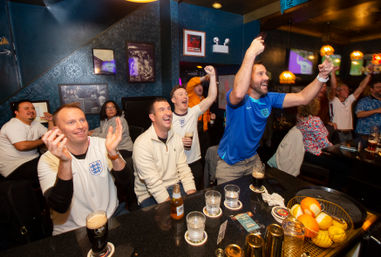 Cheering football fans at a lively pub watch party, many wearing England shirts and raising arms over a bar with pints, bottles and glasses
