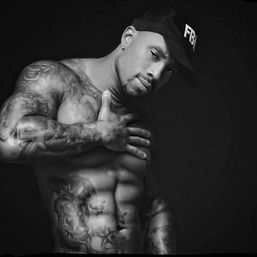 Black-and-white studio portrait of a shirtless, tattooed fitness model wearing an "FBI" cap, hand on chest and showing defined abs against a dark background.