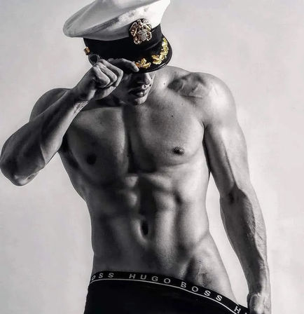 Monochrome studio photo of a shirtless, muscular fitness model tipping a white naval captain’s hat, showcasing defined abs and low-rise black briefs.