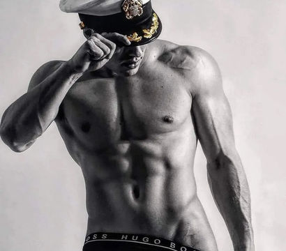 Monochrome studio photo of a shirtless, muscular fitness model tipping a white naval captain’s hat, showcasing defined abs and low-rise black briefs.